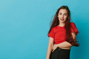Kırmızı tişörtlü ve siyah pantolonlu gülümseyen genç bir kadının portresi mavi arka planda duruyor uzun saçlar akıyor ve elleri belinde rahat ve kendinden emin bir görünüm için..