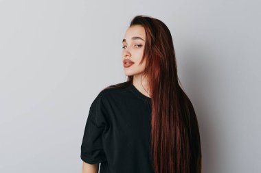 Uzun düz kırmızı saçlı, siyah bluzlu, yana bakan genç bir kadının portresi. Doğal makyajlı, modaya uygun saç stili ve sade beyaz arka plana karşı minimalist giyimli bir model..