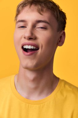 Parlak sarı tişörtlü neşeli genç adam geniş bir tebessümle gülümsüyor, sağlam bir renk arkaplanında canlı bir ifadeyle poz veriyor, iyimserlik, gençlik, sağIık ve modern moda ile ilgili kampanyalar için uygun.