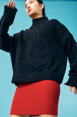 İçerde poz veren parlak kırmızı etekli, bol siyah örülü süveter giyen genç bir kadın. Minimalist arka plan, modern tarz ve giysi ayrıntıları rahat ve kendinden emin görünüm ile.