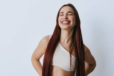 Uzun örgülü, günlük kıyafetleri olan, gözleri kapalı gülümseyen bir kadın. Neşeli ifadeli bir kadın portresi ve doğal ışıkta basit beyaz bir arkaplan..