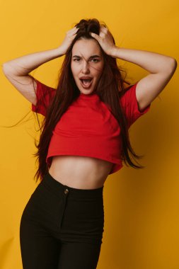 Uzun saçlı, elleri kırmızı tişörtlü ve sarı arka planda siyah pantolonlu duygusal genç bir kadın. Açık ağızlı ve moda tarzlı etkileyici bir poz..