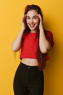 Kırmızı tişörtlü, siyah pantolonlu, uzun saçlı, gülümseyen bir kadın. Sarı arka planda yüzün yanında poz veriyor. Yaşam tarzı ve moda portresi için günlük kıyafetler giyiyor..