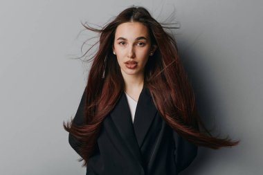 Tarafsız gri arka planda uzun saçlı ve siyah ceketli genç bir kadının portresi. Moda, güzellik ya da fotoğraf stüdyosunda yaşam tarzı için poz veren doğal görünüşlü ve dağınık saçlı bir kadın model..