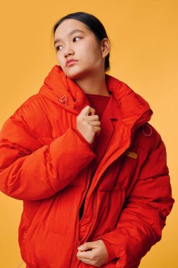 Büyük boy kırmızı kabarık ceketli Asyalı kadın model turuncu arka fonun önünde duruyor, başka tarafa bakıyor, kışlık giysiler ve modern moda, gündelik dış görünüş.