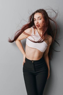 Beyaz bluzlu ve siyah pantolonlu ince kadın model, düz gri arka planda duruyor. Saçları esiyor, doğal güzelliğini gösteriyor, modern gündelik kıyafetler ve rahat ama zarif bir duruşu var..