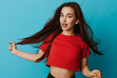 Kırmızı tişörtlü, dalgalı saçlı, mavi arka planda duran ve modern moda fotoğraf çekimlerinde rahat duruş ve moda kıyafetlerini sergileyen genç bir kadının portresi..