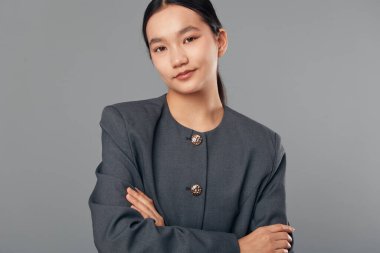 Kendine güvenen, düğmeli gri ceketli, kolları çapraz duran bir kadın. Doğal makyajlı ve ciddi yüzlü bir iş kadını portresi..