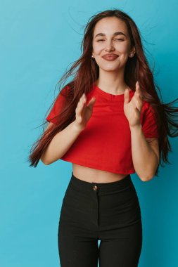 Gülümseyen genç bir kadın kırmızı gömlekli ve siyah pantolonlu ellerini çırpıyor mavi arka planda duruyor. Uzun saçlı mutlu kadın portrede neşe ve moda tarzını gösteriyor..