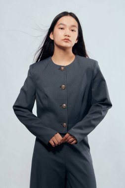 Gri takım elbiseli, metal düğmeli, elleri arkadan sıkıca bağlı poz veren bir kadın. Profesyonel ya da moda tarzında stüdyo fotoğrafında düz siyah saç ve nötr yüz ifadesi.