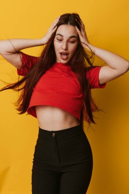 Kırmızı bluz ve siyah pantolon giyen genç bir kadının portresi açık ağızlı ve ellerinin kafasında şaşkınlık dolu duygular gösteriyor. Canlı sarı arka plan ve sıradan moda tarzıyla stüdyo çekimi.