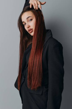 Siyah ceketli, uzun örgülü, gri arka planda duran kendine güvenen genç bir kadının portresi. Güzellik ve tarz teması için doğal ifade ve modaya uygun saç stiline sahip bir model..