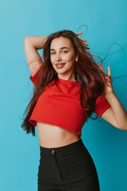 Kırmızı gömlekli ve siyah pantolonlu gülümseyen genç bir kadın. Mavi arka planda elleri saçları olan. Uzun saçlı, rahat giyimli ve stüdyo resminde rahat, kendine güvenen bir kadın model..