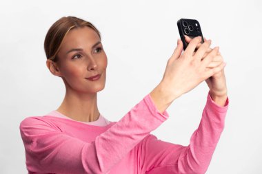 Pembe uzun kollu bluz giyen kadın akıllı telefonuyla selfie çekiyor. Beyaz arka planda parlak stüdyo ortamından emin görünüyor. Dijital iletişim ve kendini ifade etme kavramı.