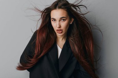 Gri arka planda dalgalı kahverengi saçlı ve siyah ceketli genç kadın portresi moda, güzellik, tarz, model ve saç konsepti için kendinden emin bir ifade sergiliyor..