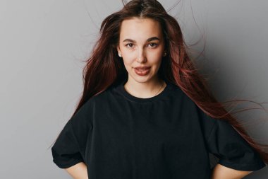 Kendine güvenen, dalgalı saçlı, siyah tişörtlü, elleri kalçasında, stüdyo ortamında gri arka planda poz veren genç bir kadının portresi..