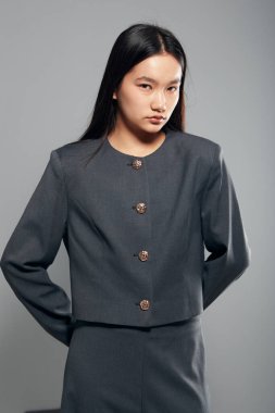 Gri takım elbiseli, düz saçlı, elleri arkada, profesyonel kıyafetli ve tarafsız stüdyo arka planında duran kendine güvenen Asyalı kadın modelin portresi..