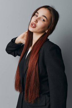 Siyah ceketli, uzun kızıl saçlı kadın içeride poz veriyor. Ciddi bir kadının stüdyo portresi. Boyuna yakın ve gri arka planda nötr bir ifade..