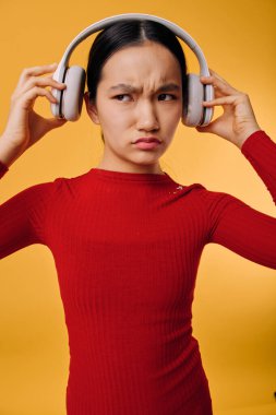 Kırmızı tişört giyen kafası karışmış bir kadın turuncu arka planda kablosuz kulaklık tutuyor. Kadın, kulaklıkları kuşkucu bir yüzle ayarlar, rahatsız edici ve ses kalitesine tepkisi belirsiz olur..