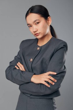 Siyah saçlı, gri iş elbisesi giyen, kolları arka planda çapraz duran kendine güvenen bir kadın manken. Ciddi bir yüzü ve stüdyo resminde düzgün bir saç stili olan genç bir profesyonel..