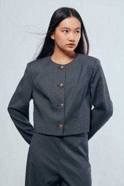 Düğmeli ceketli, elleri arkada poz veren gri takım elbiseli Asyalı kadın. Profesyonel kadın, doğal görünümlü, şirket ya da ofis çevresi için çağdaş moda..