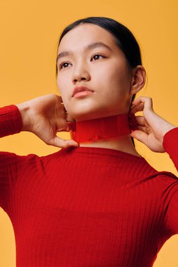 Kırmızı balıkçı yaka kazak giyen genç bir kadının portresi. İki eliyle yakasına dokunuyor. Stüdyo resmi sarı arka planda. Siyah saçlı zarif bir kadın, sakin bir ifade ve minimalist.