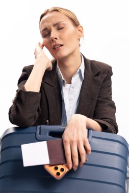 Ceketli bitkin iş kadını elinde pasaport ve telefon bulunan mavi bavula yaslanıyor. Seyahat stresi, yorgunluk ve uzun iş seyahati ya da havaalanı bekleme süresi süresince endişe kavramı.
