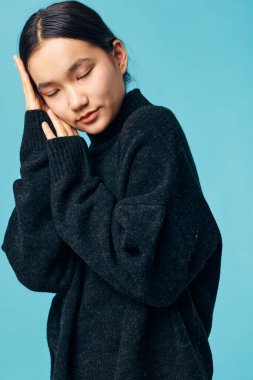 Büyük boy siyah kazak giyen kadın, elleri ve gözleri kapalı poz veriyor, mavi arka planda izole edilmiş, stüdyo resminde rahat ve huzurlu bir ruh hali sergiliyor..