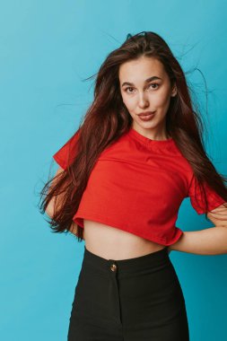 Kırmızı bluzlu, siyah pantolonlu, kendine güveni tam genç bir manken elleri kalçalarında mavi arka planda duruyor. Uzun saçlı moda portresi, günlük kıyafet ve yaşam tarzı konsepti için doğal görünüm..