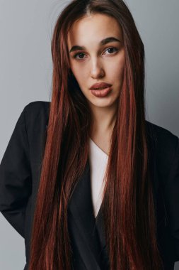 Uzun kahverengi saçlı, siyah ceketli zarif bir kadın kameraya bakıyor. Moda ya da güzellik için tarafsız arkaplanda pürüzsüz saçlı ve minimum makyajlı genç bir kadının yakın plan portresi..