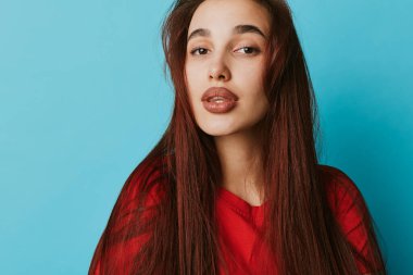 Uzun kahverengi saçlı ve kameraya bakan kırmızı elbiseli bir kadın model. Tarafsız mavi arkaplan, sıradan tarz, doğal yüz ve rahat duruş ile yakın plan portresi.