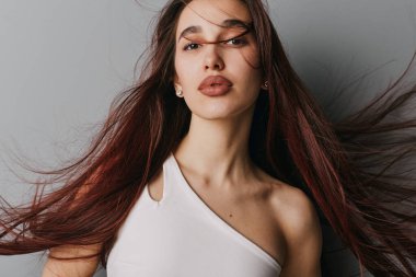 Dalgalı kahverengi saçlı, nötr makyajlı, beyaz kolsuz bluzlu, ciddi bir ifade takınan, düz gri duvarlı genç bir kadın..