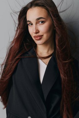 Beyaz duvarın yanında duran, uzun kahverengi saçlı, kendine güvenen genç bir kadının portresi. Doğal makyaj, rahat ifade, yumuşak gölgeli kapalı mekan ve sıradan moda tarzı..