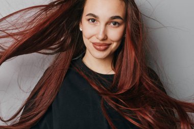 Siyah tişörtlü, parlak kızıl saçlı, doğal bir ifade ve inci küpeli genç bir kadın. Sade beyaz duvar arka planında saç hareketi olan yakın plan portresi.