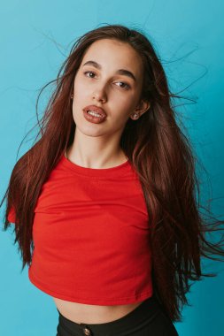 Kırmızı gömlekli, mavi arka planda uzun dalgalı saçlı genç bir kadın portresi. Doğal görünümlü ve yaşam tarzı, güzellik veya moda içeriği için rahat pozlu bir kadın modeli.