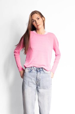 Pembe kazaklı ve açık mavi kotlu uzun düz saçlı, elleri beyaz arka planda olan genç bir kadının portresi. Günlük kıyafet ve modern moda konsepti.