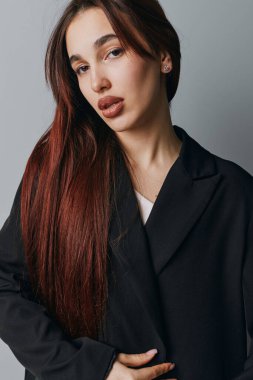 Siyah ceketli, uzun saçlı bir kadın model kameraya bakıyor. Tarafsız gri arka planda doğal görünüşü ve ince makyajı olan genç bir kadının yakın plan portresi..