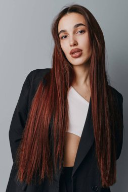 Örgülü saçlı, siyah ceketli ve beyaz bluzlu genç bir kadın stüdyoda duruyor. Modern tarz için doğal ışık ve nötr gri zemin ile yakın çekim moda çekimi.