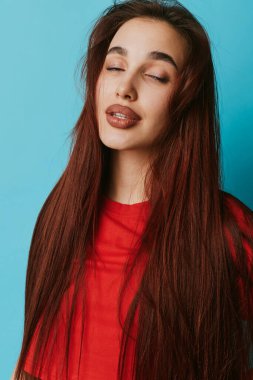 Uzun düz saçlı, kırmızı bluzlu, gözleri kapalı ve mavi arka planda sakin bir ifade takınan genç bir kadının portresi. Stüdyo çekimlerinde doğal makyaj ve dingin ruh hali..