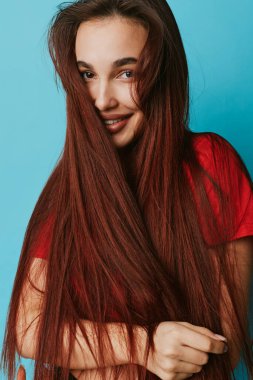 Düz, uzun saçlı, kırmızı gömlekli genç bir kadın içeride poz veriyor. Kendine güvenen bir gülümsemeyle yakın plan portresi ve yaşam tarzı ve tarz kavramı için mavi arka planda doğal bir görünüm..