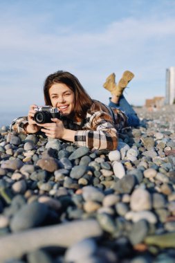 Günlük ekose ceketli kadın fotoğrafçı kayalık sahilde uzanıyor, göz hizasında kamera, açık hava fotoğrafçılığının keyfini çıkarıyor, taşlı sakin deniz manzarası, hafif esinti ve odaklanmış kompozisyon.