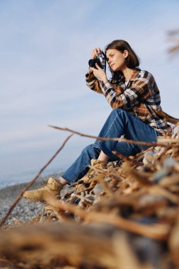 Açık hava fotoğrafçısı kamerayla deniz manzarası çekiyor, sonbahar yaprakları arasında kayaların üzerinde oturuyor, sakin bir gökyüzü, yaratıcı seyahat atmosferi, doğa ve fotoğrafçılık odaklanıyor..