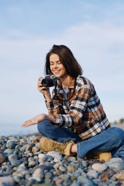 Çakıl taşı plajında sıradan bir kadın fotoğrafçı, el kamerasıyla bir kare çekmeye odaklanmış, ekose gömlek ve botlar giyiyor, doğayı kucaklıyor, seyahat titreşimleri alıyor ve açık havada rahat bir iş yapıyor.