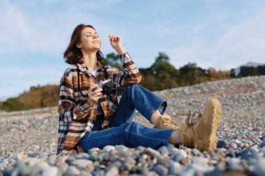 Kayalık bir plajda fotoğraf makinesi, parlak güneş ışığı, kot ve çizmeler, huzurlu bir doğa sahnesi ve günlük hayatı yakalayan sıradan bir kadın portresi..
