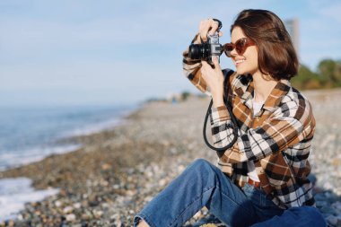 Gülümseyen bir kadın fotoğrafçı, elinde kamera, kot pantolon, ekose gömlek, güneş ışığı, deniz manzarası ve günümüzün yaratıcı açık hava görüntüleri.