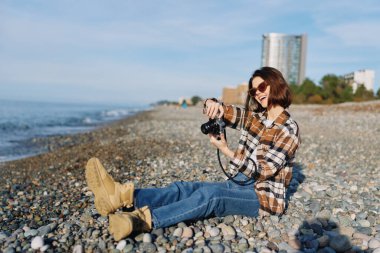 Açık hava fotoğrafçılık sahnesi. Kayalık bir sahilde sakin bir kadın nişancı. Kamera elinde. Güneşli hava, günlük kıyafet ve deniz esintisi samimi bir yolculuk anı yaratıyor..