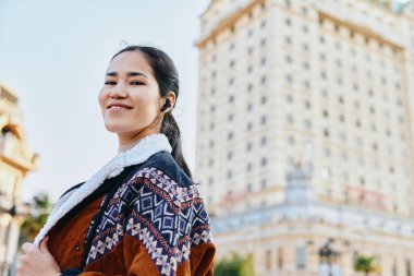 Açık hava sahnesinde kalın montlu bir kadın büyük mimariye, şehir sokaklarına ve seyahat havasına karşı gülümsüyor.