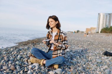 Kareli tişört giyen sıradan fotoğrafçı kadın kayalık sahilde oturuyor, elinde bir kamera, dalgalar yükseldikçe gülümsüyor, deniz kenarındaki manzara fotoğrafçılığı seansı.