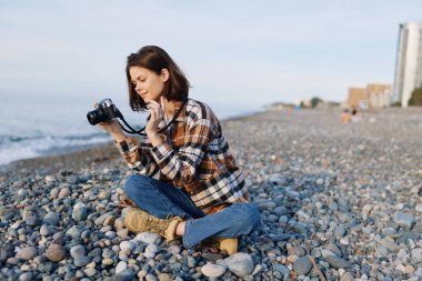 Kayalık bir sahilde sıradan bir fotoğrafçı, elinde kamera, ekose gömlek ve botlarla çömelmiş bir kadın, ufka odaklanıyor, kıyı dokusu ve nazik gelgit, sakin bir açık hava yolculuğu anı yaratıyor.