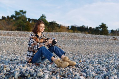 Dışarıda kameralı bir kadın, kumsal boyunca çakıl taşlarına oturmuş gündelik giysiler, doğa odaklı fotoğrafçılık ve hobi, huzurlu bir an ve temiz gökyüzü altında manzaralı bir manzara.